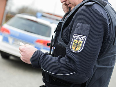 Ein Polizist in uniform, der ein Smartphone in der Hand hält, im Hintergrund ein Polizeiwagen.