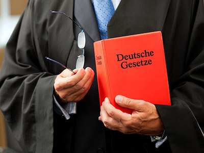 Ein Richter hält ein rotes Buch mit der Aufschrift "Deutsche Gesetze" und eine Brille in der Hand.
